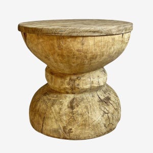 May include: A rustic, wooden stool with a natural, light brown finish. The stool has a round top, a bulbous midsection, and a rounded base. The wood grain is visible throughout the piece, adding to its natural aesthetic. The stool is a unique piece of furniture.