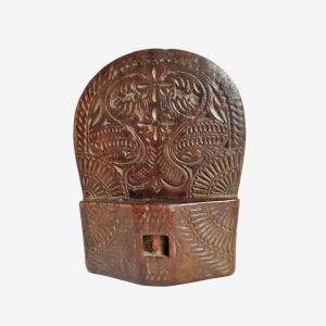 May include: A hand-carved wooden object with intricate floral and leaf patterns. The dark brown piece has a rectangular base and a curved top, featuring detailed relief carvings. The design includes symmetrical floral motifs and sunburst-like patterns.