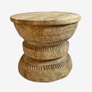May include: A light brown, wooden stool with a round top and a carved, hourglass-shaped base. The wood grain is visible throughout the piece, and the base features decorative carved lines. The stool is a rustic, decorative item.