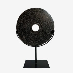 May include: A dark, circular stone sculpture with a central hole, mounted on a black metal stand. The stone has a textured surface and is displayed against a white background. The sculpture is a decorative art piece.