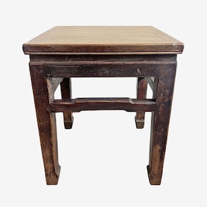 May include: A small, square wooden stool with a light-colored top and dark brown legs. The legs are rectangular and have a slight curve at the bottom. The stool is approximately 12 inches tall.