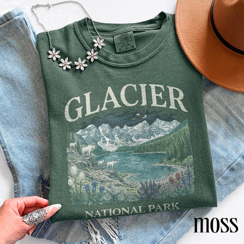 Puede incluir: Una sudadera verde musgo con la inscripci&oacute;n "GLACIER NATIONAL PARK" y una ilustraci&oacute;n de monta&ntilde;as, un lago y fauna. Tambi&eacute;n se ven un collar plateado con colgantes en forma de flor y un sombrero marr&oacute;n.