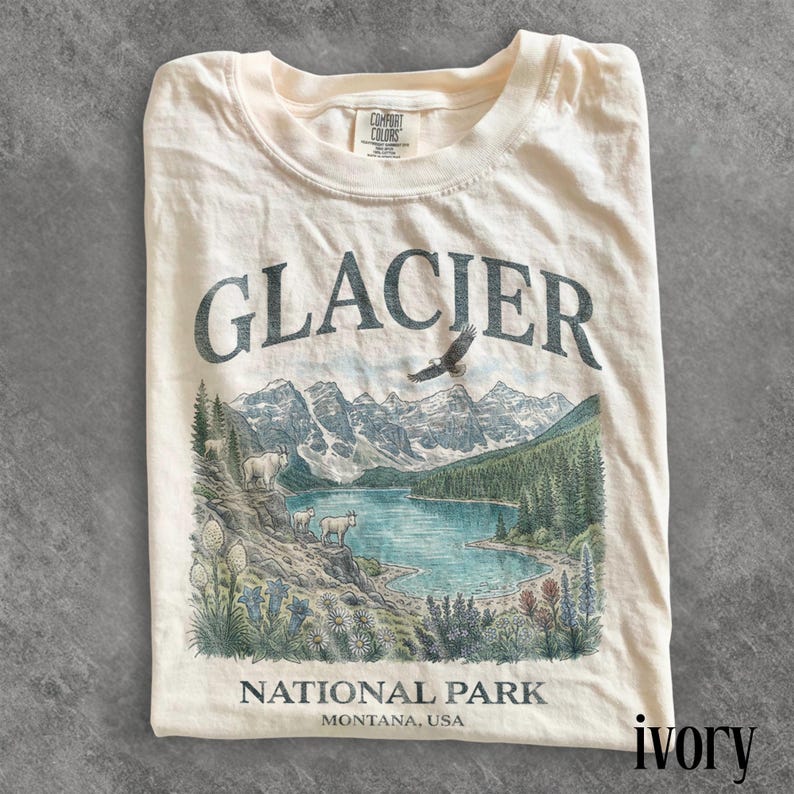 Puede incluir: Camiseta de color marfil con una ilustraci&oacute;n del Parque Nacional Glacier. El dise&ntilde;o incluye la palabra "GLACIER" sobre una escena de lago con monta&ntilde;as, &aacute;rboles y fauna. El texto "NATIONAL PARK MONTANA, USA" est&aacute; impreso debajo.