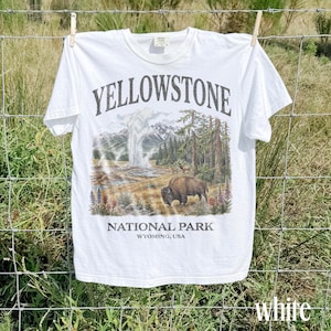 May include: White t-shirt featuring a Yellowstone National Park graphic. The design includes a geyser, mountains, trees, and a bison. The text "YELLOWSTONE NATIONAL PARK WYOMING, USA" is printed above and below the graphic.