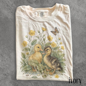 Peut inclure: T-shirt ivoire avec une illustration détaillée de deux canetons au milieu de fleurs jaunes et de feuillage vert, avec un papillon. Le t-shirt a un col rond et la marque "Comfort Colors" est visible. Le mot "ivoire" est imprimé en bas.