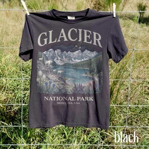 Puede incluir: Camiseta negra con una ilustraci&oacute;n del Parque Nacional Glacier, Montana, EE. UU. El dise&ntilde;o incluye un lago, monta&ntilde;as y el texto "GLACIER NATIONAL PARK". La camiseta est&aacute; colgada en un tendedero.