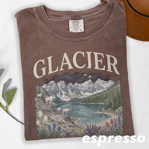 Puede incluir: Camiseta color espresso con la palabra "GLACIER" en letras grandes de color blanquecino. El gr&aacute;fico representa una escena de lago de monta&ntilde;a con monta&ntilde;as, &aacute;rboles y fauna. La camiseta es de la marca Comfort Colors.