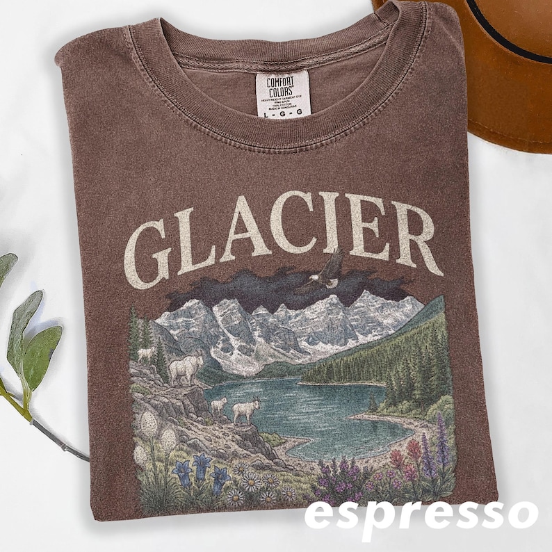 Puede incluir: Camiseta color espresso con la palabra "GLACIER" en letras grandes de color blanquecino. El gr&aacute;fico representa una escena de lago de monta&ntilde;a con monta&ntilde;as, &aacute;rboles y fauna. La camiseta es de la marca Comfort Colors.