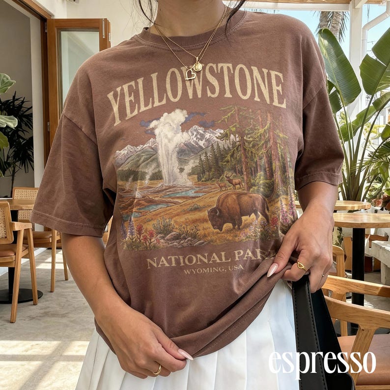 May include: Brown t-shirt with a Yellowstone National Park graphic. The design features a landscape with a geyser, mountains, trees, and a bison. The text "YELLOWSTONE NATIONAL PARK WYOMING, USA" is printed on the shirt.
