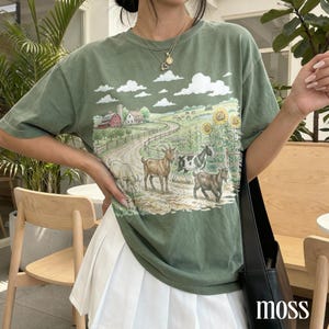 Könnte beinhalten: Olivgrünes T-Shirt mit einer ländlichen Szene: Ziegen, roter Bauernhof, Sonnenblumen und ein Weg. Das T-Shirt hat eine lockere Passform und kurze Ärmel. Das Wort "moss" ist unten rechts in Weiß gedruckt.
