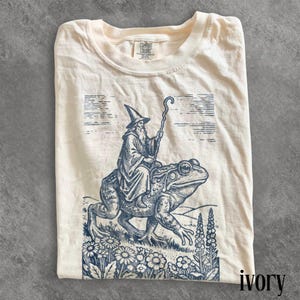 Puede incluir: Camiseta color marfil con una ilustración azul de un mago montado en una rana, con un bastón en la mano. El diseño incluye elementos florales y paisajísticos. La camiseta es de Comfort Colors. La palabra 'ivory' está impresa.