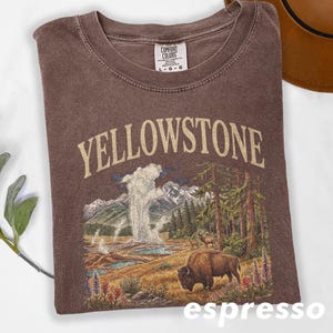May include: Espresso-colored t-shirt with a graphic of Yellowstone National Park. The design includes the word "YELLOWSTONE" in a tan font, a geyser, mountains, trees, and a bison. The shirt has a crew neck and a Comfort Colors tag.