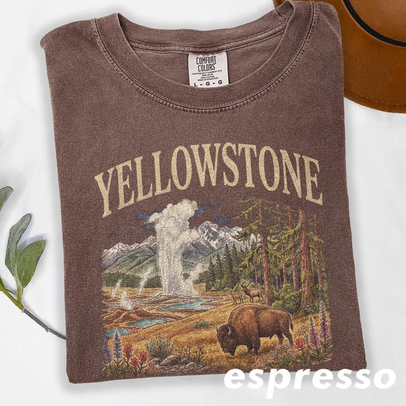 May include: Espresso-colored t-shirt with a graphic of Yellowstone National Park. The design includes the word "YELLOWSTONE" in a tan font, a geyser, mountains, trees, and a bison. The shirt has a crew neck and a Comfort Colors tag.
