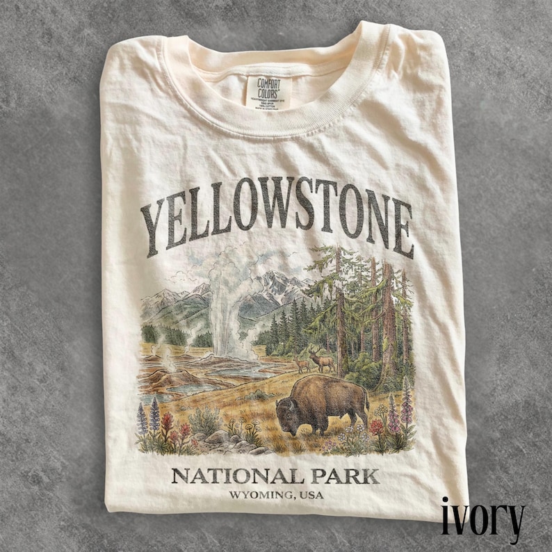 May include: Ivory-colored t-shirt featuring a graphic of Yellowstone National Park. The design includes the text "YELLOWSTONE NATIONAL PARK WYOMING, USA" with an illustration of a geyser, bison, and trees.
