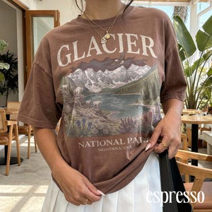Puede incluir: Camiseta marr&oacute;n con un gr&aacute;fico de un lago de monta&ntilde;a y la palabra "GLACIER" en blanco. Tambi&eacute;n es visible el texto "NATIONAL PARK MONTANA, USA". La camiseta se combina con una falda blanca.