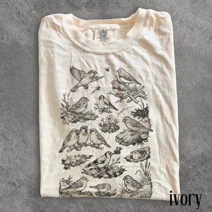 May include: Ivory-colored t-shirt with a detailed black and white illustration of various birds perched on branches and foliage. The shirt has a crew neck and the brand name "Comfort Colors" is visible. The word "ivory" is printed at the bottom right.