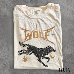 May include: Ivory-colored t-shirt featuring a black wolf graphic with the word "WOLF" in gold lettering. The design includes a star and crescent moon. The shirt is folded and has a Comfort Colors tag.