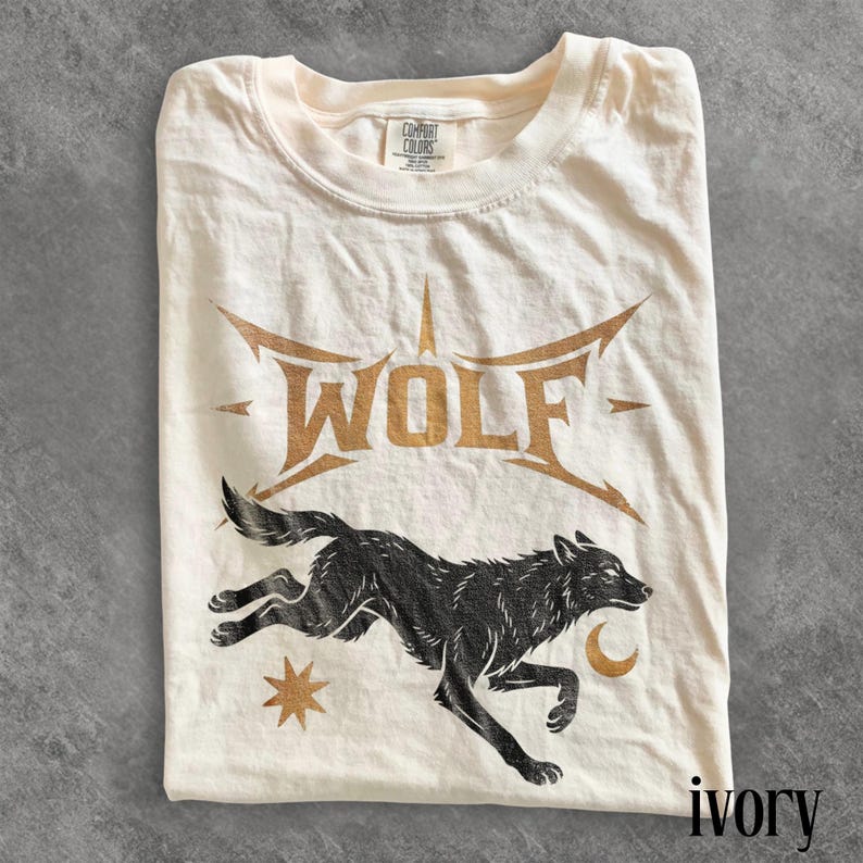 May include: Ivory-colored t-shirt featuring a black wolf graphic with the word "WOLF" in gold lettering. The design includes a star and crescent moon. The shirt is folded and has a Comfort Colors tag.