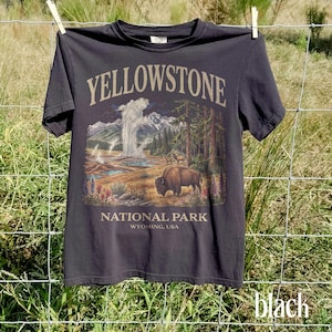 May include: Black t-shirt featuring a Yellowstone National Park design. The graphic includes a bison, geyser, mountains, and trees. The text "YELLOWSTONE NATIONAL PARK WYOMING, USA" is printed on the shirt.