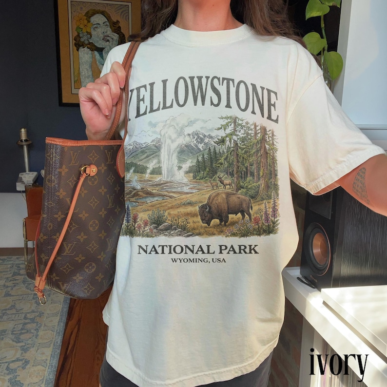 May include: Ivory-colored t-shirt featuring a Yellowstone National Park graphic. The design includes a geyser, bison, trees, and mountains. The text "YELLOWSTONE NATIONAL PARK WYOMING, USA" is printed on the shirt.