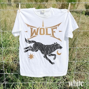 May include: White t-shirt with a graphic of a running black wolf, a star, and a crescent moon. The word "WOLF" is printed in gold above the wolf. The word "white" is printed in white at the bottom right.