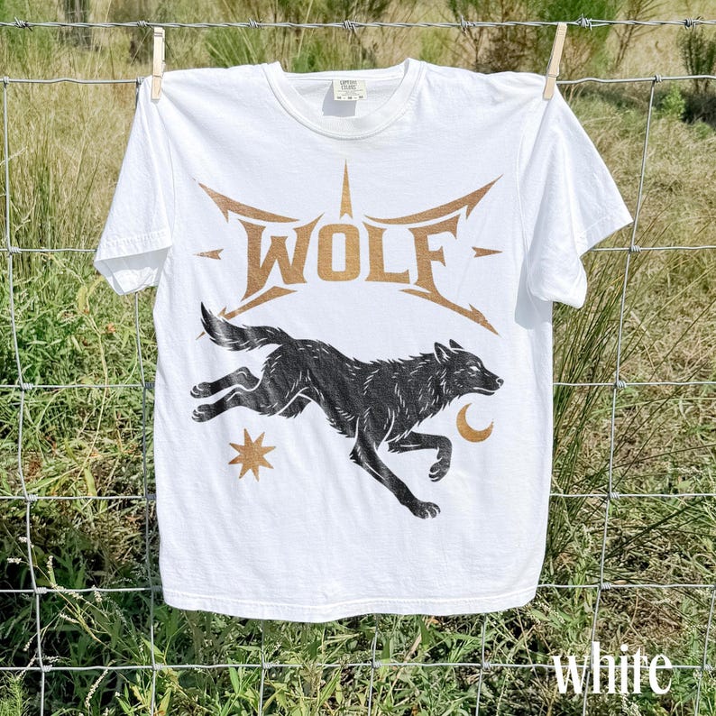 May include: White t-shirt with a graphic of a running black wolf, a star, and a crescent moon. The word "WOLF" is printed in gold above the wolf. The word "white" is printed in white at the bottom right.