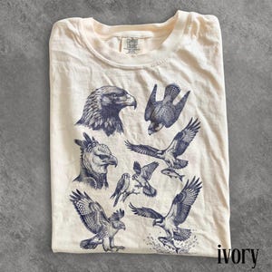 Könnte beinhalten: Elfenbeinfarbenes T-Shirt mit marineblauen Illustrationen von Raubvögeln. Das Shirt hat den Text "COMFORT COLORS" auf dem Etikett und "ivory" unten rechts. Das Design zeigt Adler, Falken und andere Greifvögel.