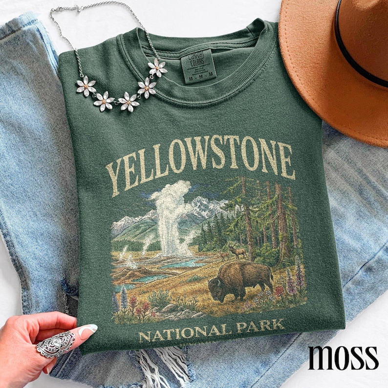 May include: A moss green sweatshirt featuring a Yellowstone National Park graphic with a bison, geyser, and mountain scene. The shirt has a crew neck and short sleeves. A silver necklace with flower-shaped pendants is on top of the shirt.