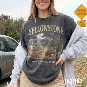 May include: Dark gray t-shirt featuring a Yellowstone National Park graphic. The design includes a geyser, mountains, trees, and a bison. The text "YELLOWSTONE NATIONAL PARK WYOMING, USA" is printed on the shirt.