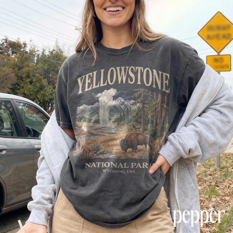 May include: Dark gray t-shirt featuring a Yellowstone National Park graphic. The design includes a geyser, mountains, trees, and a bison. The text "YELLOWSTONE NATIONAL PARK WYOMING, USA" is printed on the shirt.