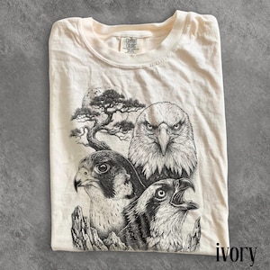 Könnte beinhalten: Elfenbeinfarbenes T-Shirt mit einer detaillierten Schwarz-Weiß-Illustration von drei Raubvögeln, darunter ein Adler und ein Falke. Das Design beinhaltet einen Baum und einen Vollmond. Das Shirt ist von Comfort Colors.