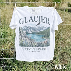 Puede incluir: Camiseta blanca con una ilustraci&oacute;n paisaj&iacute;stica del Parque Nacional Glacier, Montana, EE. UU. El dise&ntilde;o incluye un lago, monta&ntilde;as, &aacute;rboles y un &aacute;guila, con la palabra "GLACIER" arriba y "NATIONAL PARK" abajo.