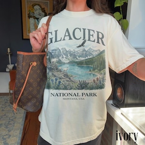Puede incluir: Camiseta de color marfil con un dise&ntilde;o gr&aacute;fico del Parque Nacional Glacier. El dise&ntilde;o presenta un lago, monta&ntilde;as, &aacute;rboles y el texto "GLACIER NATIONAL PARK MONTANA, USA". La camiseta es de manga corta y cuello redondo.