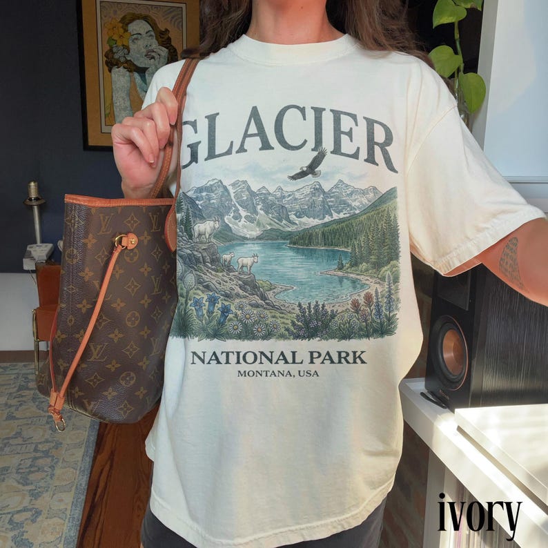 Puede incluir: Camiseta de color marfil con un dise&ntilde;o gr&aacute;fico del Parque Nacional Glacier. El dise&ntilde;o presenta un lago, monta&ntilde;as, &aacute;rboles y el texto "GLACIER NATIONAL PARK MONTANA, USA". La camiseta es de manga corta y cuello redondo.