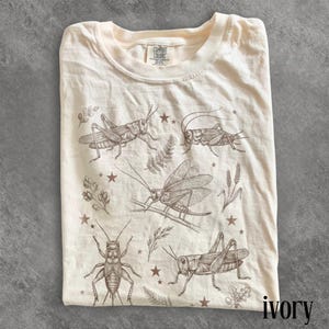 May include: Ivory-colored t-shirt featuring a detailed insect illustration. The design includes various grasshoppers, stars, and botanical elements. The shirt is made of a soft material and has a classic crew neck. The brand label "Comfort Colors" is visible.