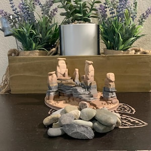 May include: A small desert landscape diorama featuring layered rock formations in shades of tan, grey, and brown. A pile of smooth, grey and tan river rocks sits in front. The scene is set against a backdrop of potted plants and a wooden box.