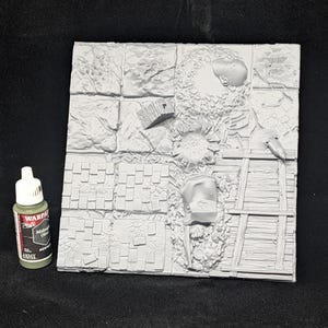 May include: A gray, textured miniature terrain tile set with various surface details, including stone, wood, and rubble. A bottle of Midnight Olive acrylic paint is next to the tile set.