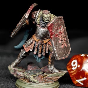 May include: A miniature orc warrior figurine, posed with a shield and weapon. The orc is clad in detailed armor with a fur collar, and the shield has a blood-splattered design. A red and gold die with the number 9 is in the foreground.