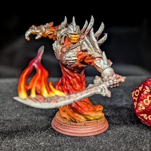 May include: A miniature fantasy figure of a fiery demon, wielding a sword. The figure is painted in shades of red, orange, and silver, with flames emanating from its base and sword. The figure is on a round base, and a red die is in the background.