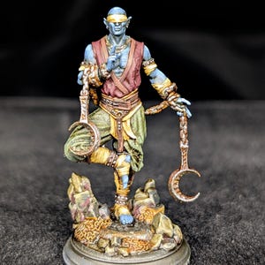 May include: A miniature figurine of a blue-skinned humanoid figure with a blindfold, holding two crescent-shaped weapons. The figure is dressed in a maroon vest, green pants, and gold accents, standing on a rocky base. The figure is approximately 3 inches tall.