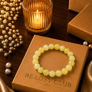 May include: A circular bracelet made of smooth, pale yellow beads resting on a square tan box with the text BEADSY CLUB embossed in gold. The beads have a polished, semi-translucent finish and are arranged in a continuous loop on the box surface.