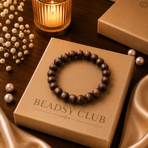 May include: A circular bracelet made of dark brown wooden beads resting on a square tan cardboard box. The box features the text BEADSY CLUB printed in a dark brown font. The beads have a visible wood grain texture and a smooth, rounded finish.