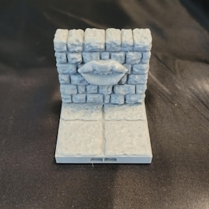 May include: A gray, 3D-printed miniature of a brick wall with a pair of lips in the center. The wall sits on a rectangular base with two square tiles. The model is designed for tabletop gaming or display.