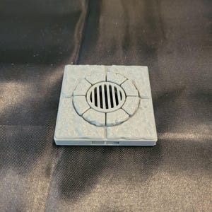 May include: A square, light gray stone-textured tile featuring a circular drain cover in the center. The drain cover consists of a series of vertical slats surrounded by a ring of eight individual stone segments, all set within a flat square base.