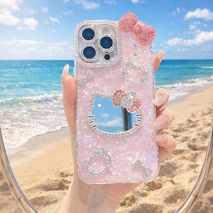 May include: A pink phone case featuring a Hello Kitty design, adorned with rhinestones and a bow. The case has a reflective surface and is set against a beach background.