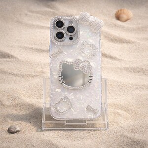 May include: Clear phone case with a white pearlescent design and sparkling rhinestone accents. Features a large Hello Kitty outline, smaller Hello Kitty outlines, and a bow. The camera lens is surrounded by rhinestones. The case is on a clear stand.