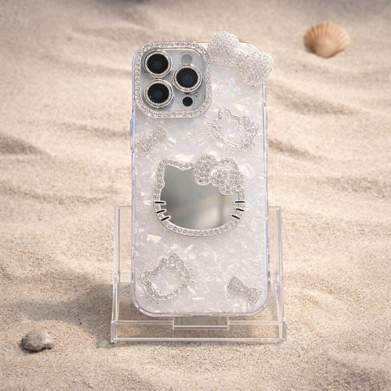 May include: Clear phone case with a white pearlescent design and sparkling rhinestone accents. Features a large Hello Kitty outline, smaller Hello Kitty outlines, and a bow. The camera lens is surrounded by rhinestones. The case is on a clear stand.