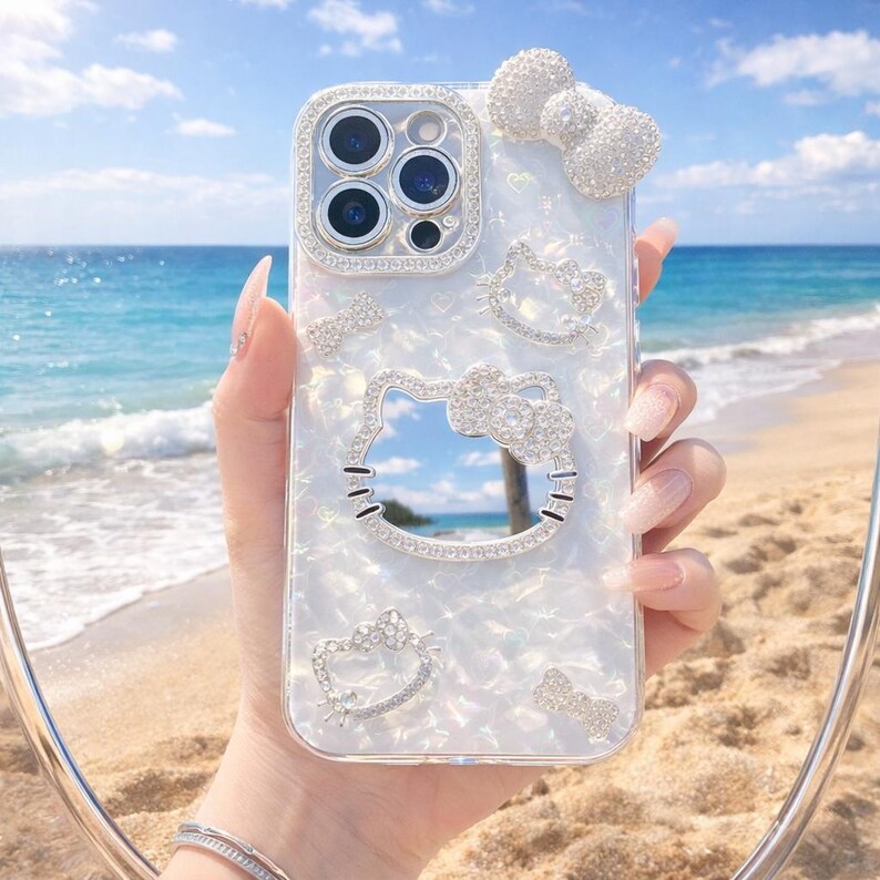 May include: A clear phone case with a pearlescent finish, decorated with sparkling rhinestones. The case features a bow, heart, and Hello Kitty designs. The phone's camera lenses are visible, surrounded by a rhinestone border. The background shows a beach and ocean.