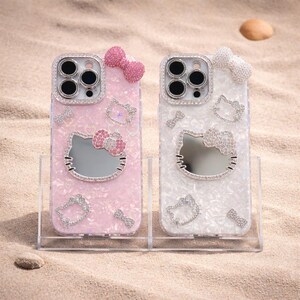 May include: Two phone cases with a Hello Kitty design. One is pink, the other white, both with a pearlised finish. Each case features a mirror Hello Kitty face, bow, and rhinestone accents around the camera and the Hello Kitty face.