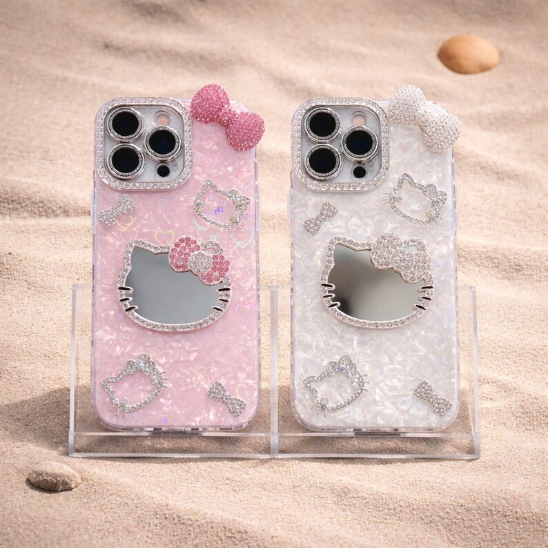 May include: Two phone cases with a Hello Kitty design. One is pink, the other white, both with a pearlised finish. Each case features a mirror Hello Kitty face, bow, and rhinestone accents around the camera and the Hello Kitty face.